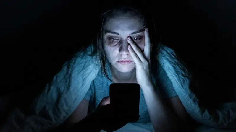Persona iluminada por la luz azul del móvil en la oscuridad, con expresión de cansancio y aislamiento